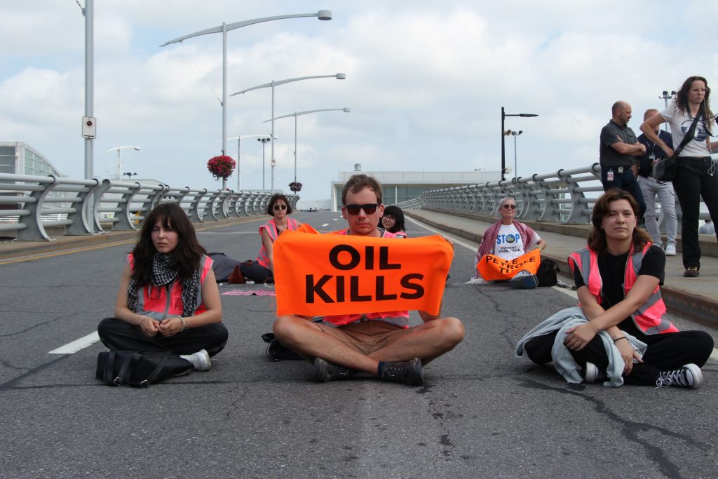 People sitting down on the street wearing bright orange shirts and holding bright orange sign reading "OIL KILLS"